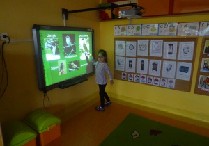 Dziewczynka stoi przy tablicy w prawej uniesionej dłoni trzyma wskaźnik skierowany na pokrzewkę.
